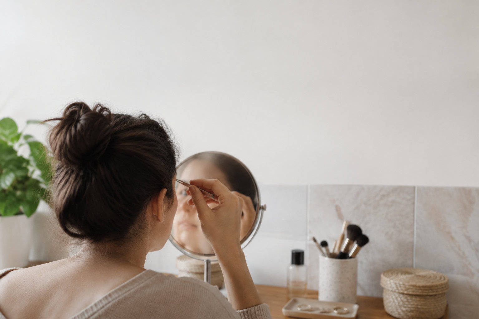 Une femme se regarde dans un miroir et utilise une pince à épiler pour entretenir la forme de ses sourcils.